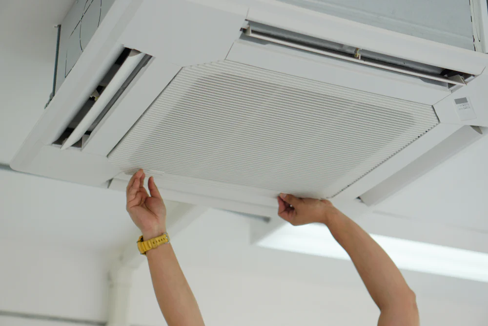 A person’s hands, wearing a yellow wristwatch, are opening or adjusting a ceiling-mounted air conditioning unit panel in TX, possibly preparing for an ac replacement San Antonio service.