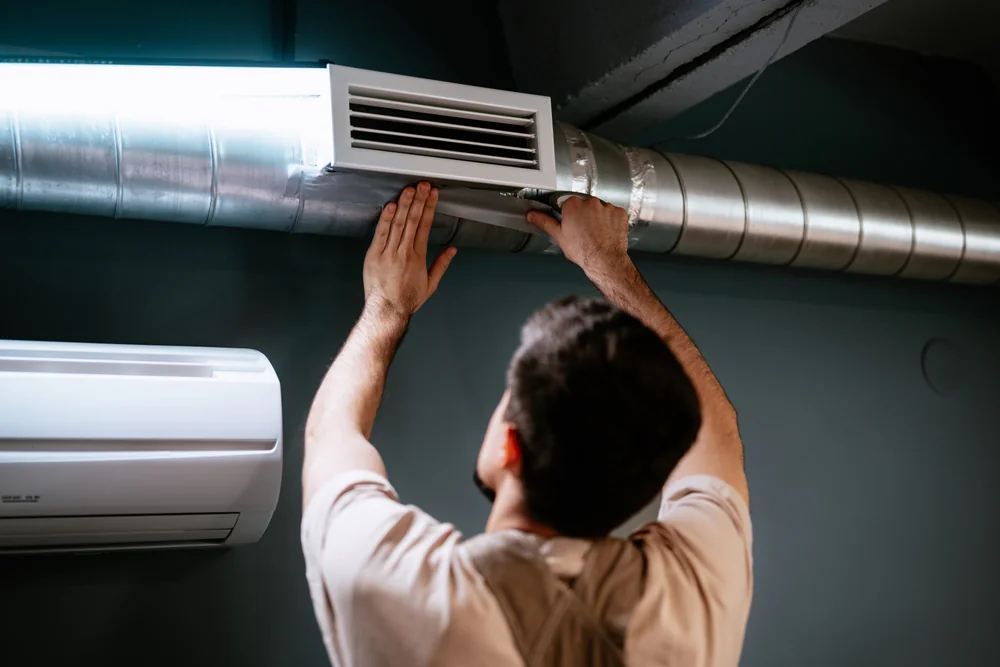 A person adjusts or installs a vent cover on a metal air duct attached to the ceiling in TX, with a wall-mounted air conditioning unit visible nearby, possibly during an ac replacement San Antonio project.