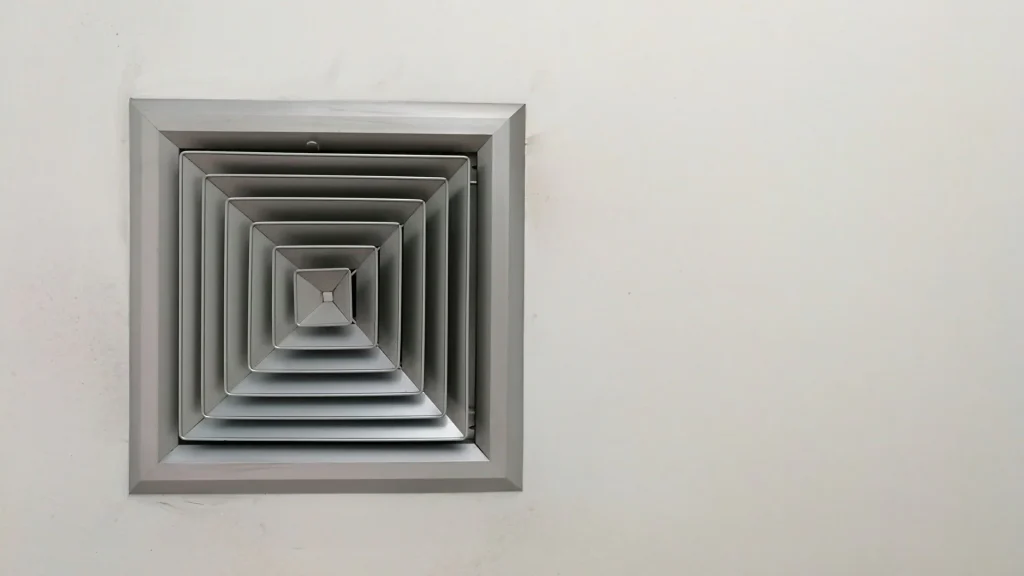 A square metal air vent with concentric layers is mounted on a plain white wall—its geometric, symmetrical pattern suggests recent ac replacement in San Antonio, TX, appearing clean and unused.