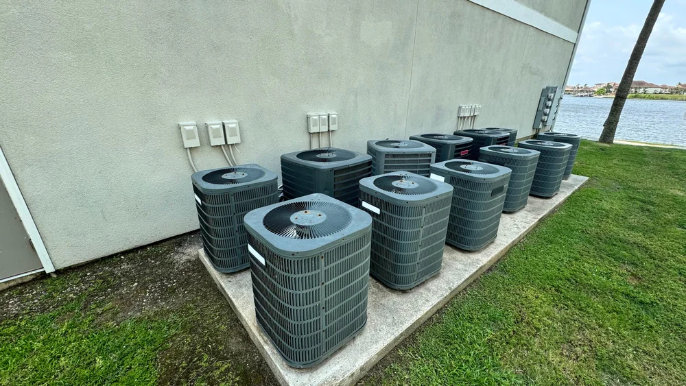 Ten outdoor air conditioning units are lined up on a concrete pad next to a beige building in Bexar County, TX. Green grass surrounds the area, with a body of water and homes visible in the background—an ideal spot for AC replacement.