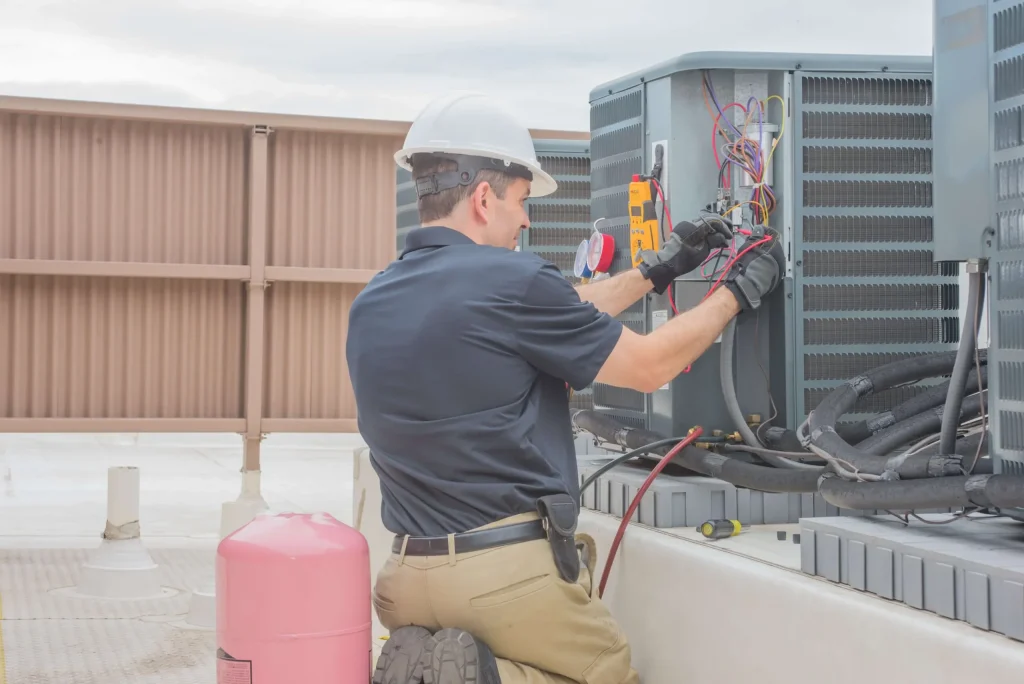 A technician wearing a hard hat and gloves kneels on a rooftop in San Antonio, TX, inspecting and testing the wiring of an air conditioning unit—an essential step before any ac replacement. Tools and equipment are visible nearby.