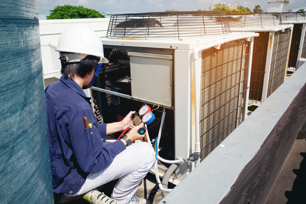 A technician wearing a white hard hat and blue uniform checks air conditioning units on a rooftop in San Antonio, TX, using pressure gauges and tools—an expert in AC replacement and maintenance.