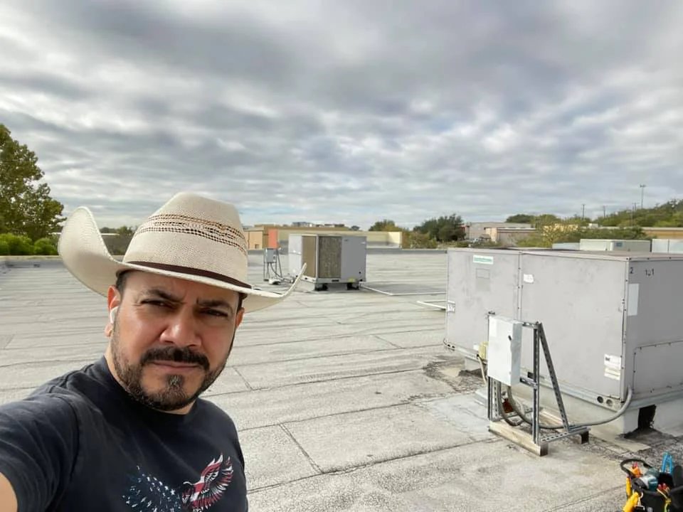 A man in a cowboy hat takes a selfie on a flat rooftop with HVAC units, perhaps during an AC replacement San Antonio project. Cloudy skies loom above, while trees and distant buildings frame the Texas (TX) backdrop.