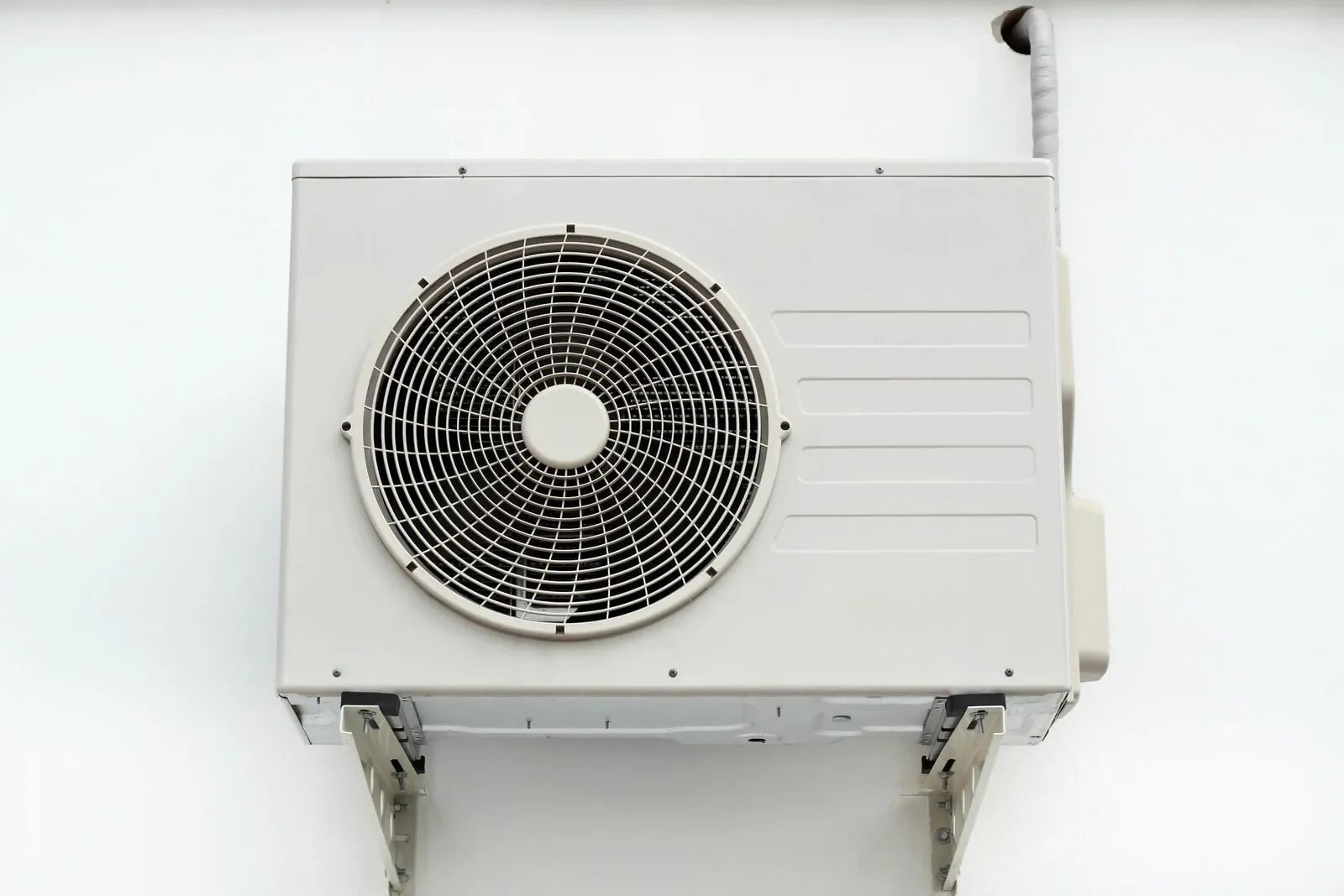 A white outdoor air conditioning unit mounted on a wall in TX, featuring a large circular fan grille and pipes extending into the wall above&mdash;ideal for AC replacement in San Antonio.