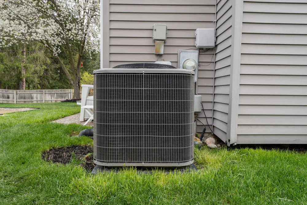A large outdoor air conditioning unit sits on a concrete pad next to the exterior wall of a gray house in Bexar County, TX, surrounded by green grass and landscaping—ideal after a recent ac replacement.