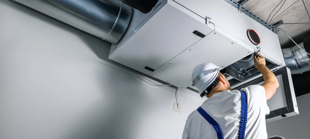 A worker in a white hard hat and overalls performs maintenance on an industrial HVAC ventilation unit, adjusting a component with one hand&mdash;typical of professional AC replacement in San Antonio, TX.