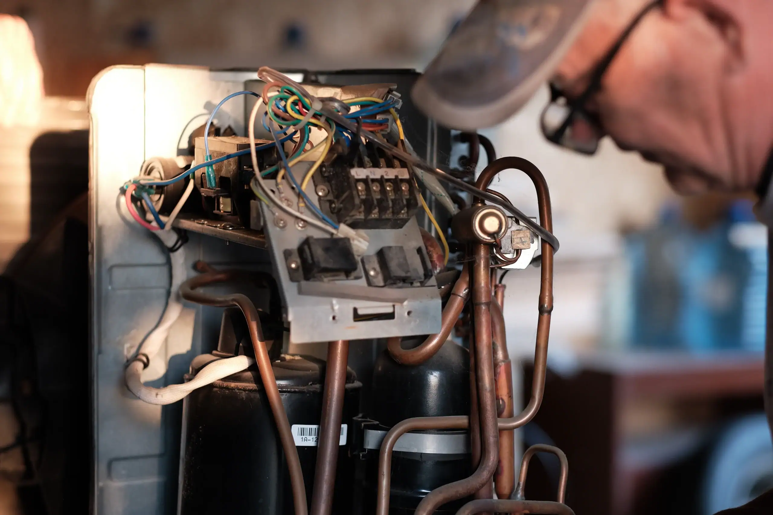A person wearing glasses and a cap inspects the exposed wiring and internal components of an electrical appliance, possibly a refrigerator, in a workshop setting.