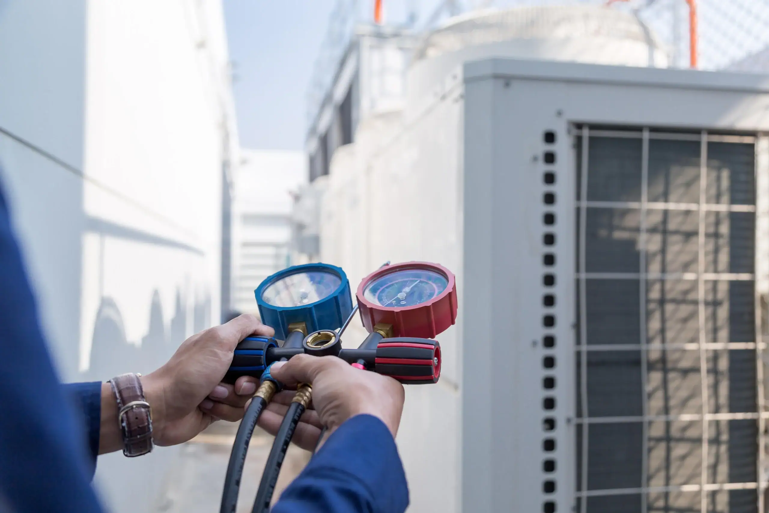 A technician holds a manifold gauge set to check the pressure on an outdoor air conditioning unit, with buildings and equipment visible in the background.