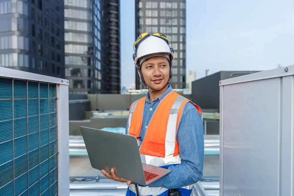 A construction worker wearing a safety helmet and orange vest holds a laptop while standing between two large equipment units on a rooftop, with modern buildings in the background.