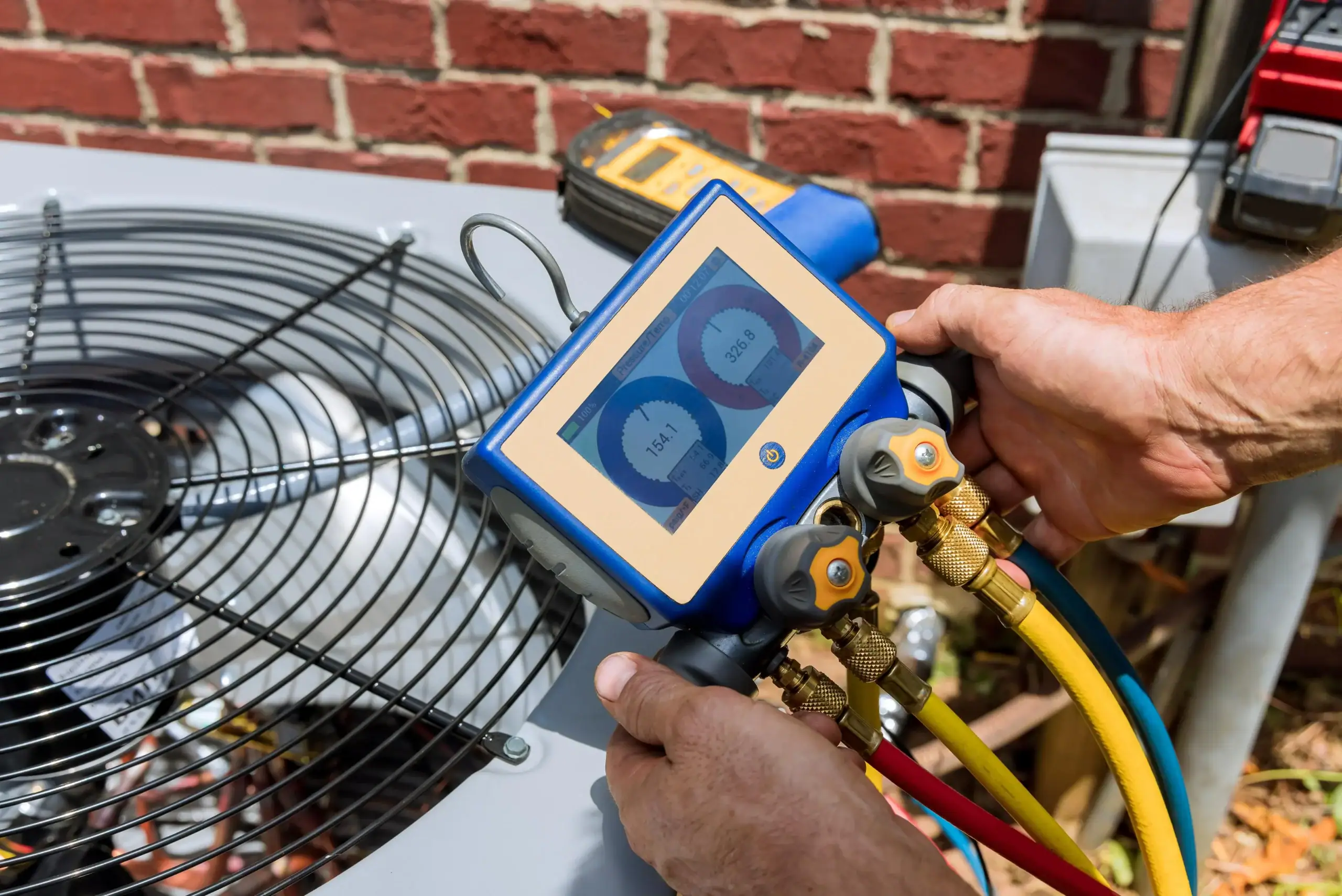 A technician uses a digital gauge manifold to check an air conditioning unit outdoors. The device displays pressure readings, and colorful hoses are connected to the AC system near a brick wall.