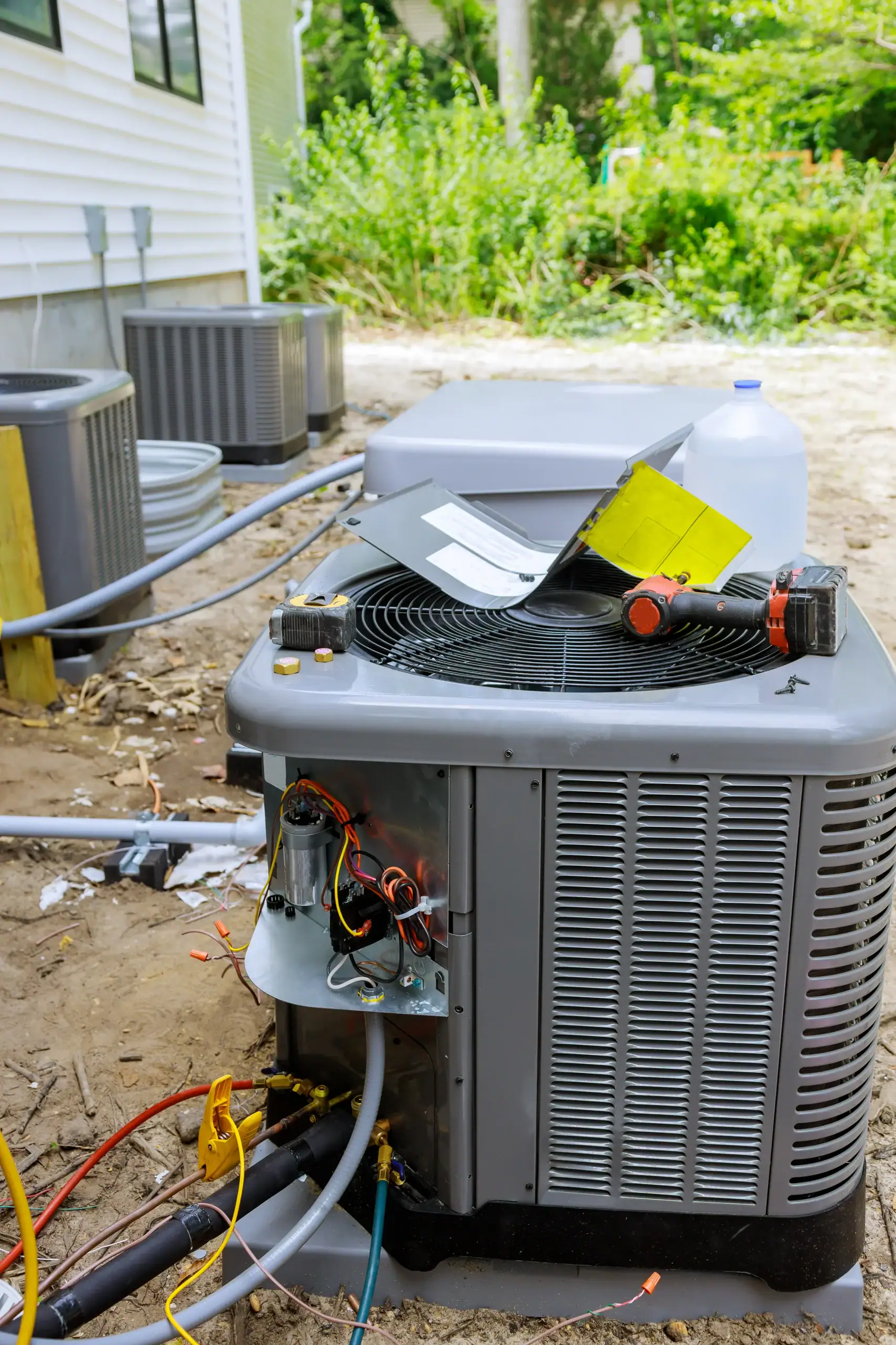 An outdoor air conditioning unit with its cover removed, exposing electrical wiring and components, sits on bare ground near a house. Nearby are tools, papers, a plastic water jug, and additional AC units.
