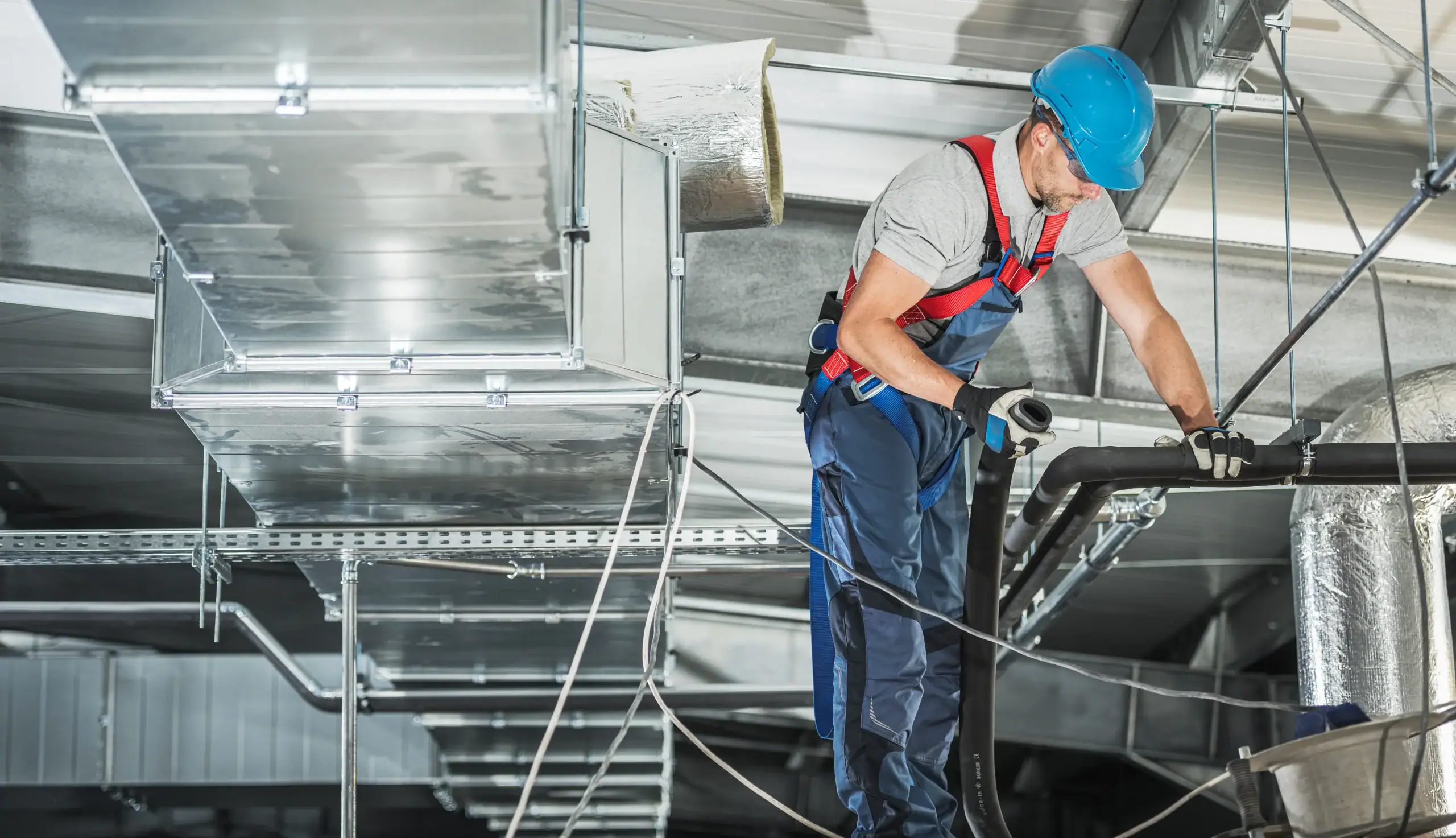 A worker in safety gear and a blue hard hat stands on a platform, installing or repairing air ducts and pipes in an industrial building's ceiling.