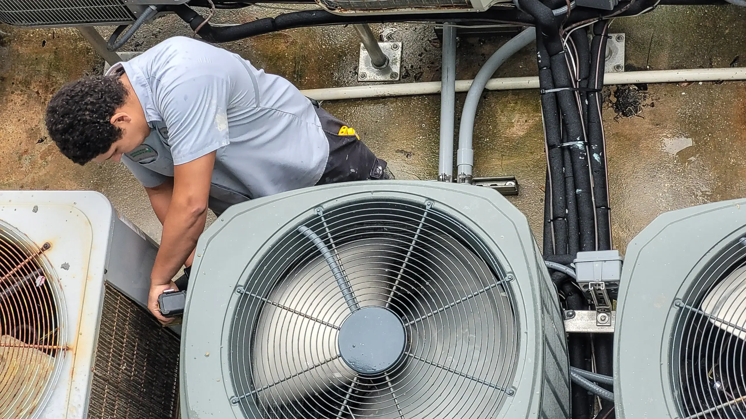 A person in a gray shirt repairs or inspects an outdoor air conditioning unit, surrounded by several large metal HVAC systems and electrical wires.