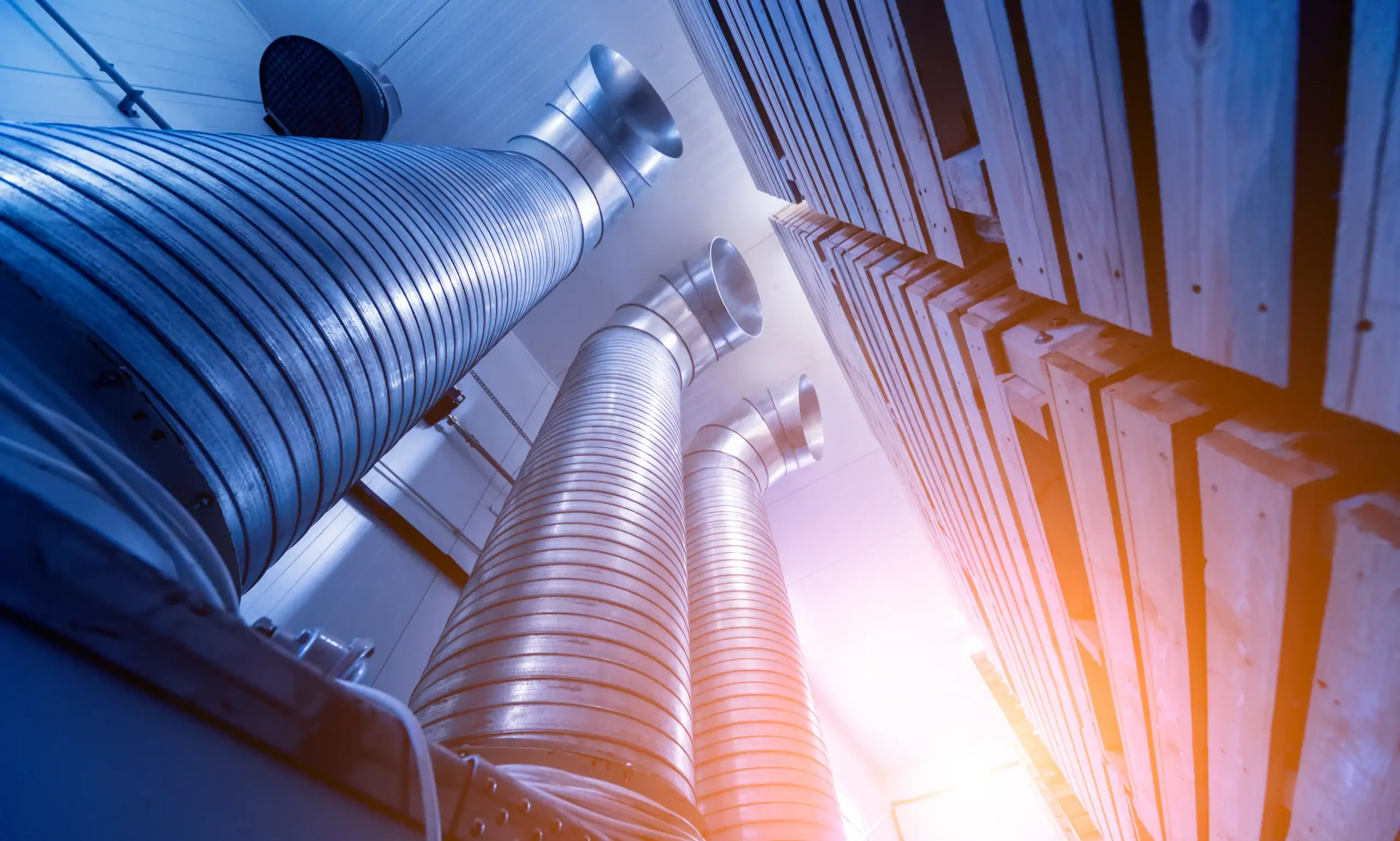 Industrial air ducts made of metal extending vertically alongside large wooden crates, with bright sunlight filtering into the room, creating a modern, industrial atmosphere.