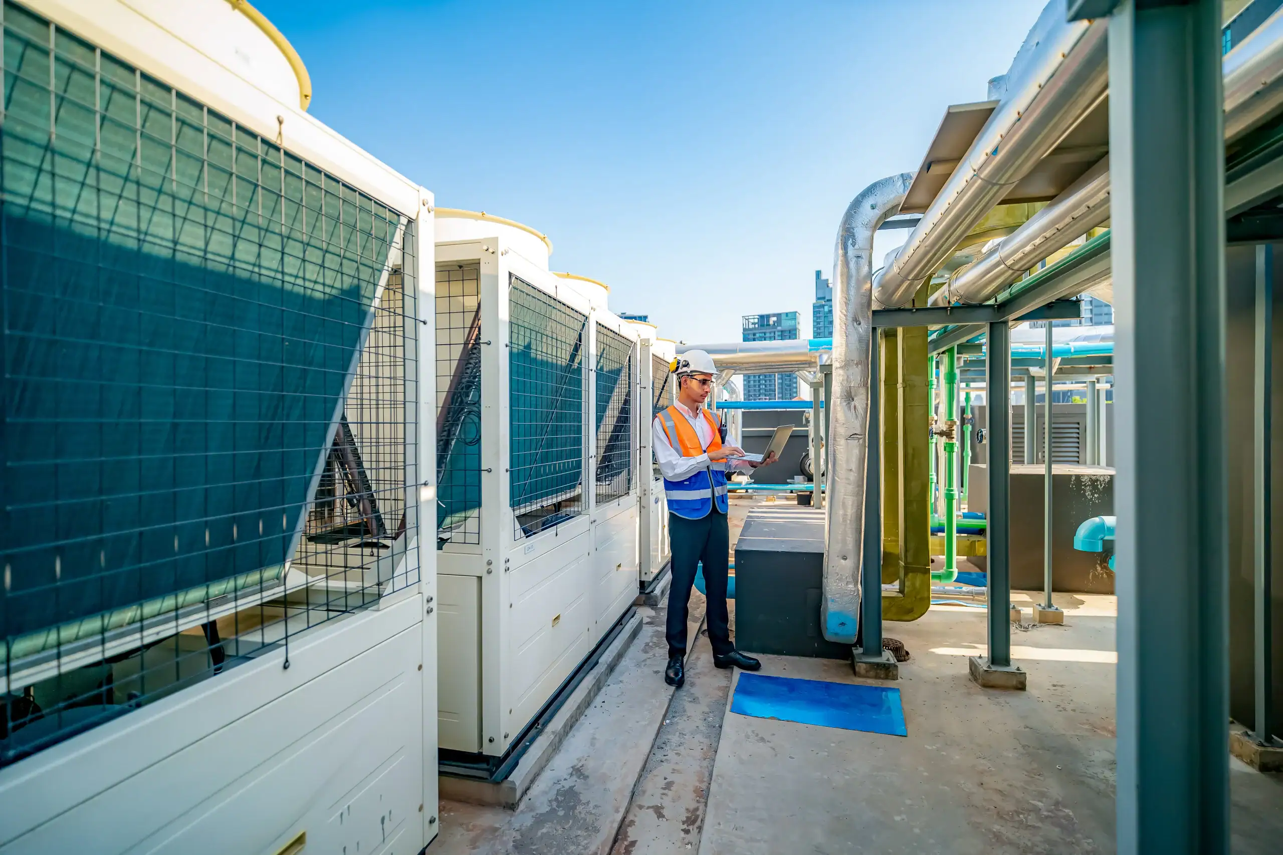 An engineer wearing a safety vest and helmet inspects industrial HVAC equipment on a rooftop, holding a laptop and standing between large units and metal pipes under a clear blue sky.