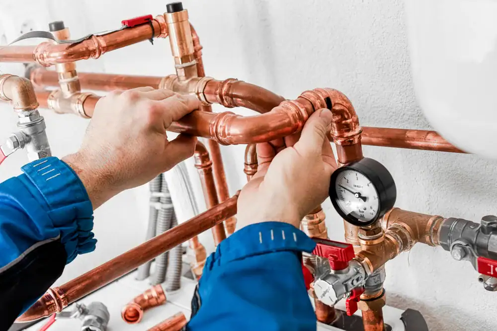 A plumber wearing a blue uniform adjusts copper pipes with a pressure gauge attached, working on a complex plumbing system against a white wall.