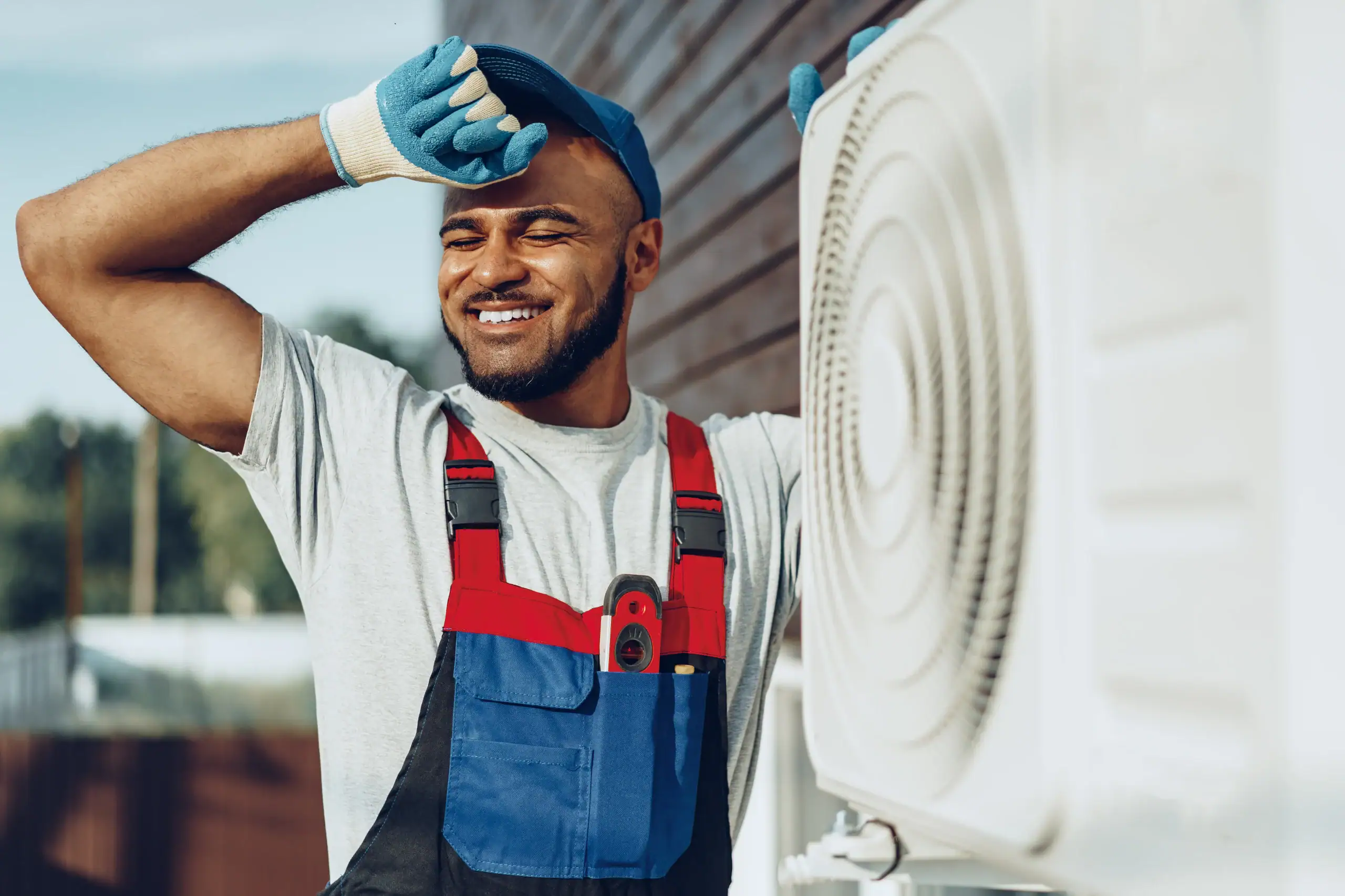 A smiling technician wearing gloves and overalls wipes his forehead while standing next to an outdoor air conditioning unit attached to a building.