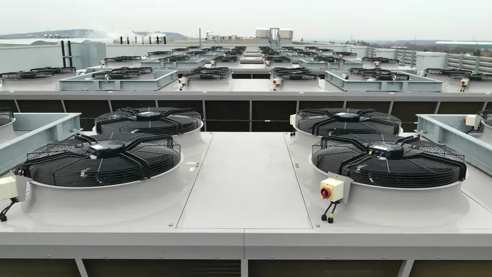 Rows of large industrial rooftop cooling fans and ventilation units are arranged in parallel lines atop a modern building, with an overcast sky and distant landscape visible in the background.