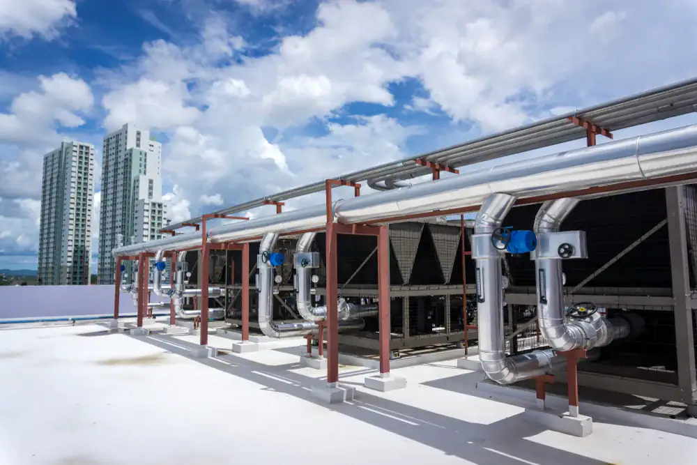Large rooftop HVAC units with metal pipes and valves are installed on a building. Tall residential or office buildings are visible in the background under a partly cloudy sky.
