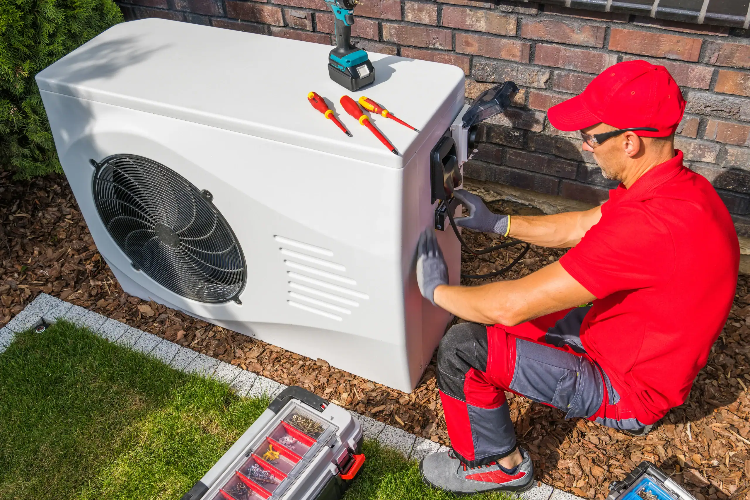 Technician in red uniform and cap repairs an outdoor heat pump unit next to a brick wall, using tools from a toolbox and several screwdrivers placed on top of the unit.