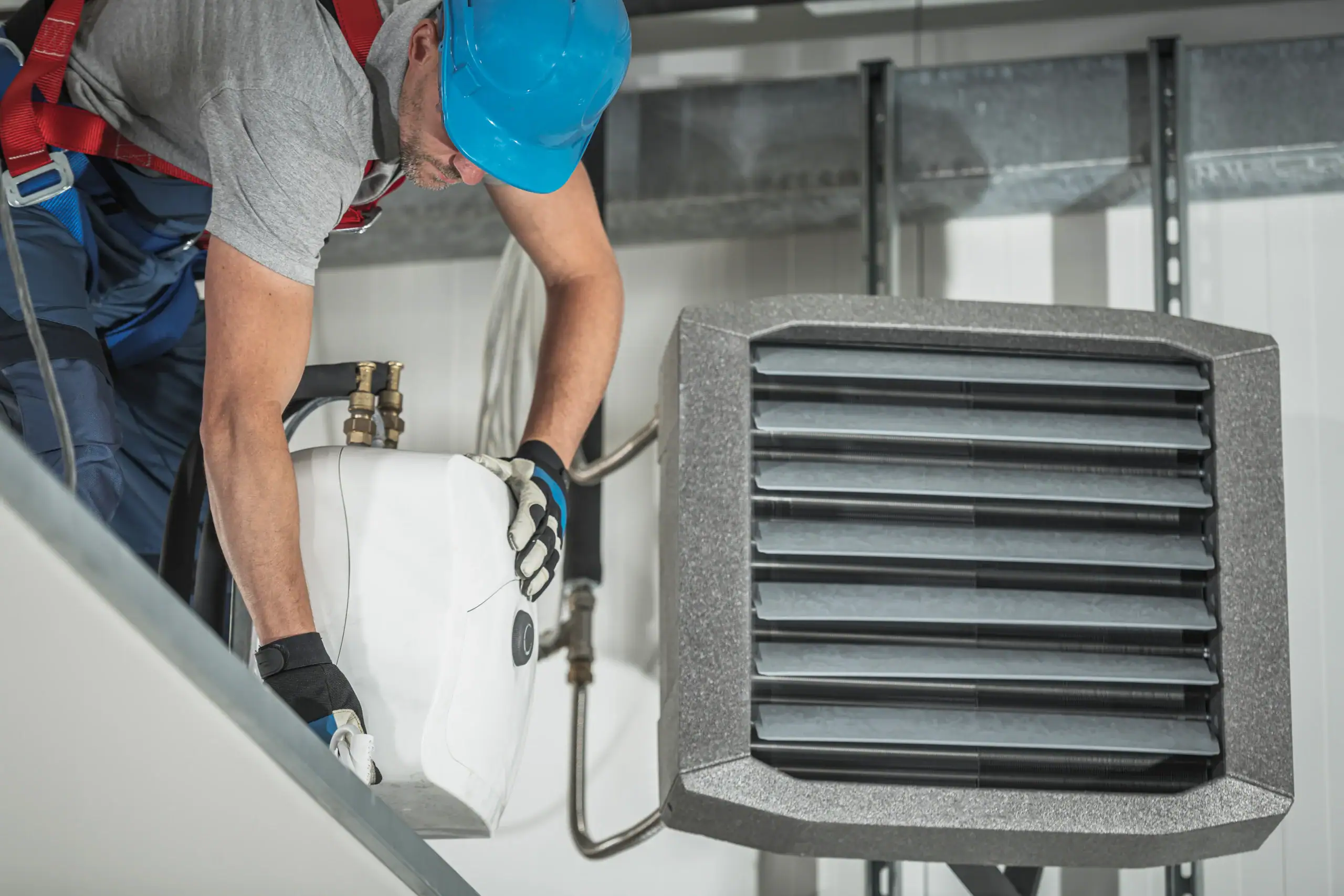 A worker in a blue hard hat and safety harness fills or services an industrial air conditioning or heating unit with a liquid container indoors. The unit features large black horizontal vents.
