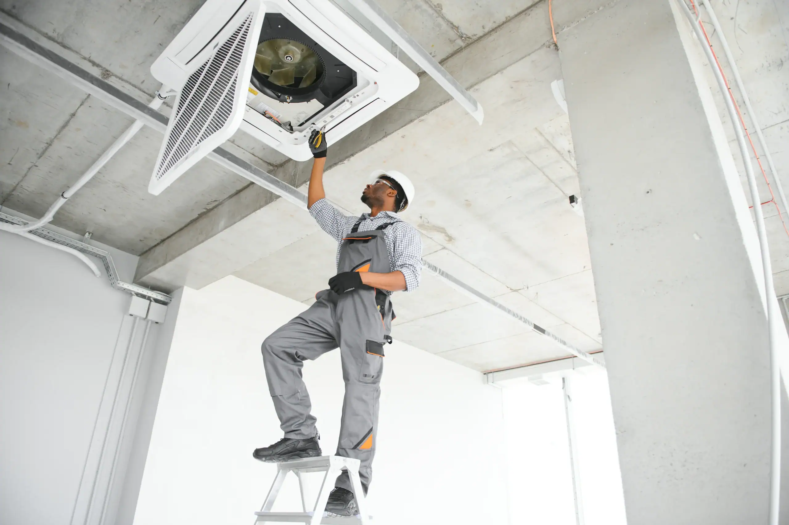 A maintenance worker in uniform and a hard hat stands on a ladder, inspecting or repairing a ceiling-mounted air conditioning unit in a modern, unfinished room with concrete ceilings.