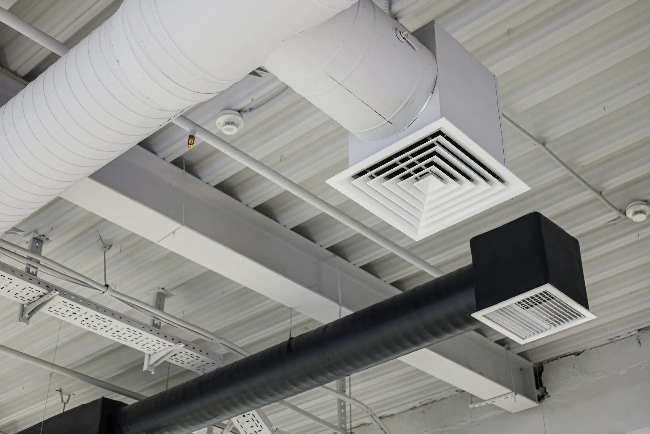 Ceiling view of an industrial building showing exposed air ducts, including a white square air vent and a black rectangular vent, with metal beams and conduit trays.