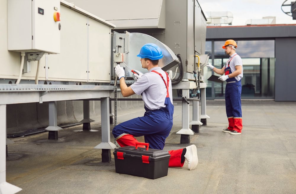 Two workers in uniforms and hard hats perform maintenance on rooftop HVAC units; one kneels using tools while the other stands in the background; a toolbox sits on the ground nearby.