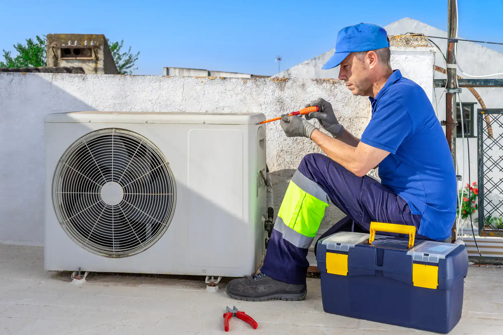 A technician wearing a blue uniform and cap uses a screwdriver to repair an outdoor air conditioning unit. Providing HVAC services in San Antonio, TX, he works on a rooftop with a toolbox and pliers beside him.