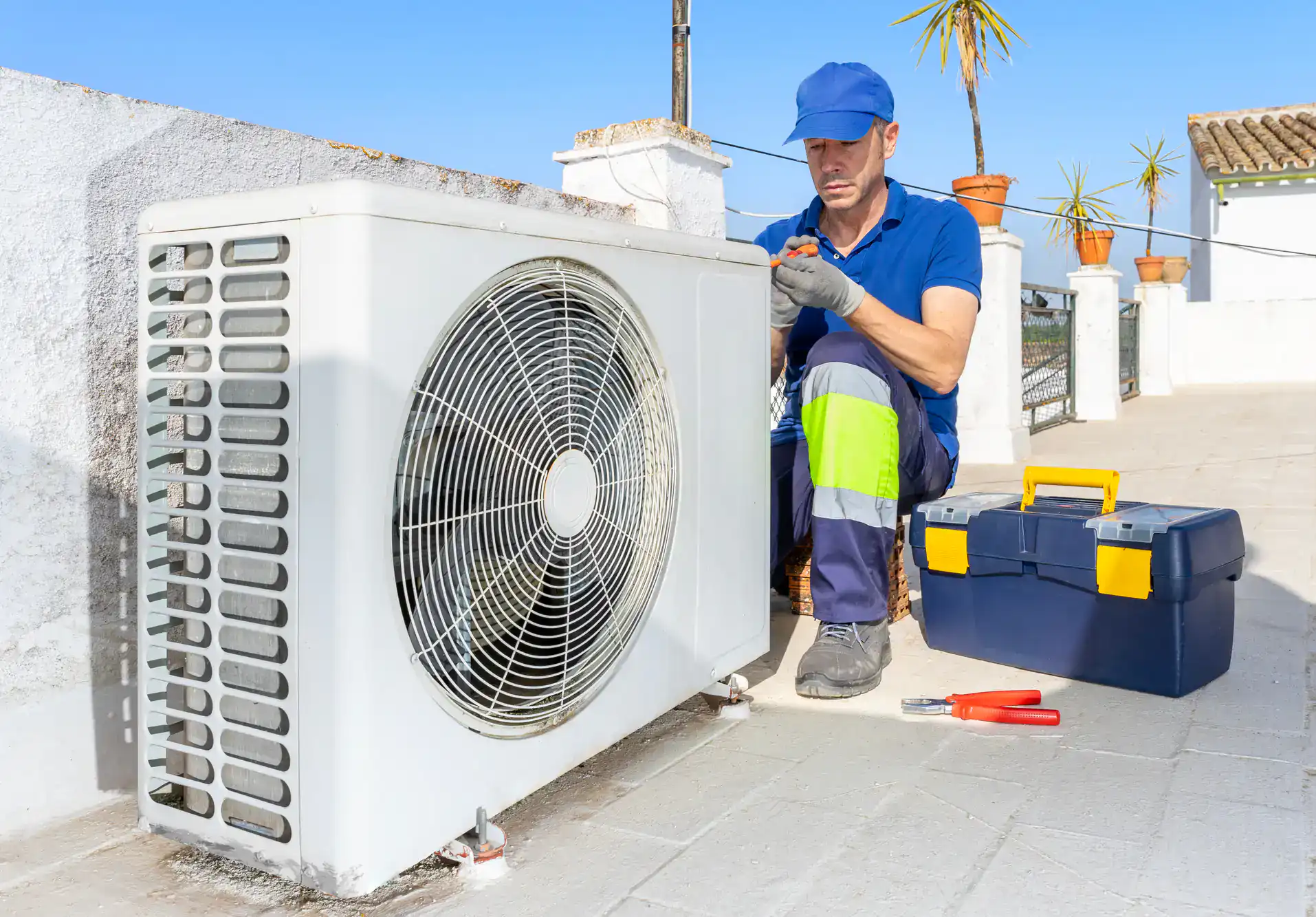 A technician in a blue uniform and cap repairs an outdoor air conditioning unit on a sunny TX rooftop patio. Nearby are a toolbox and pliers, with potted plants in the background&mdash;representing HVAC Services in San Antonio.