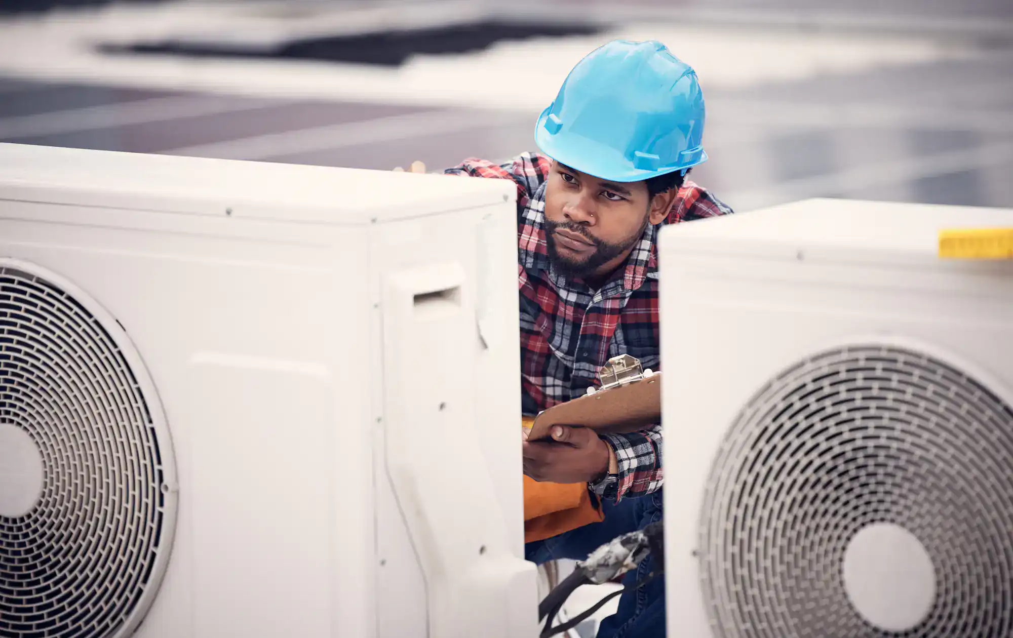 A worker wearing a blue hard hat and plaid shirt inspects outdoor air conditioning units on a rooftop, holding a clipboard and looking closely at the equipment&mdash;providing expert HVAC services in San Antonio, TX.